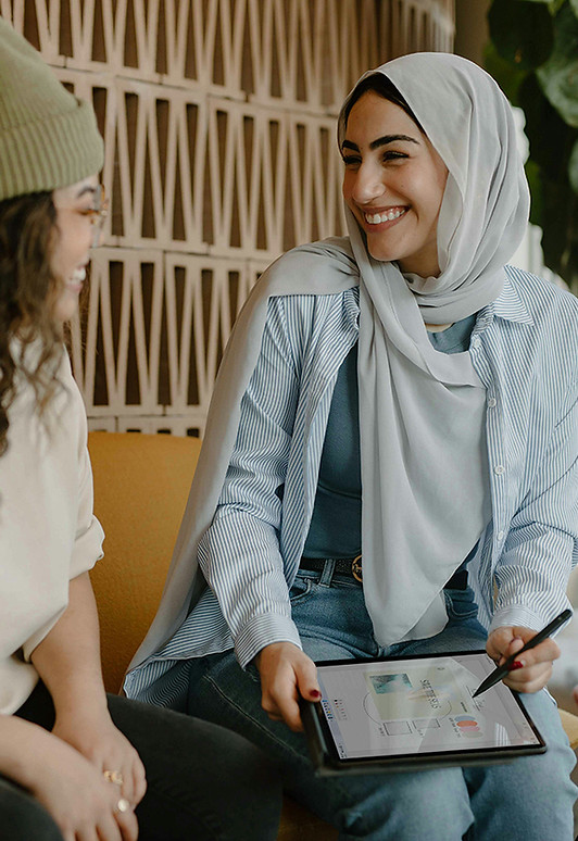 Two women conversing, one of them is wearing a head covering and holding an iPAD